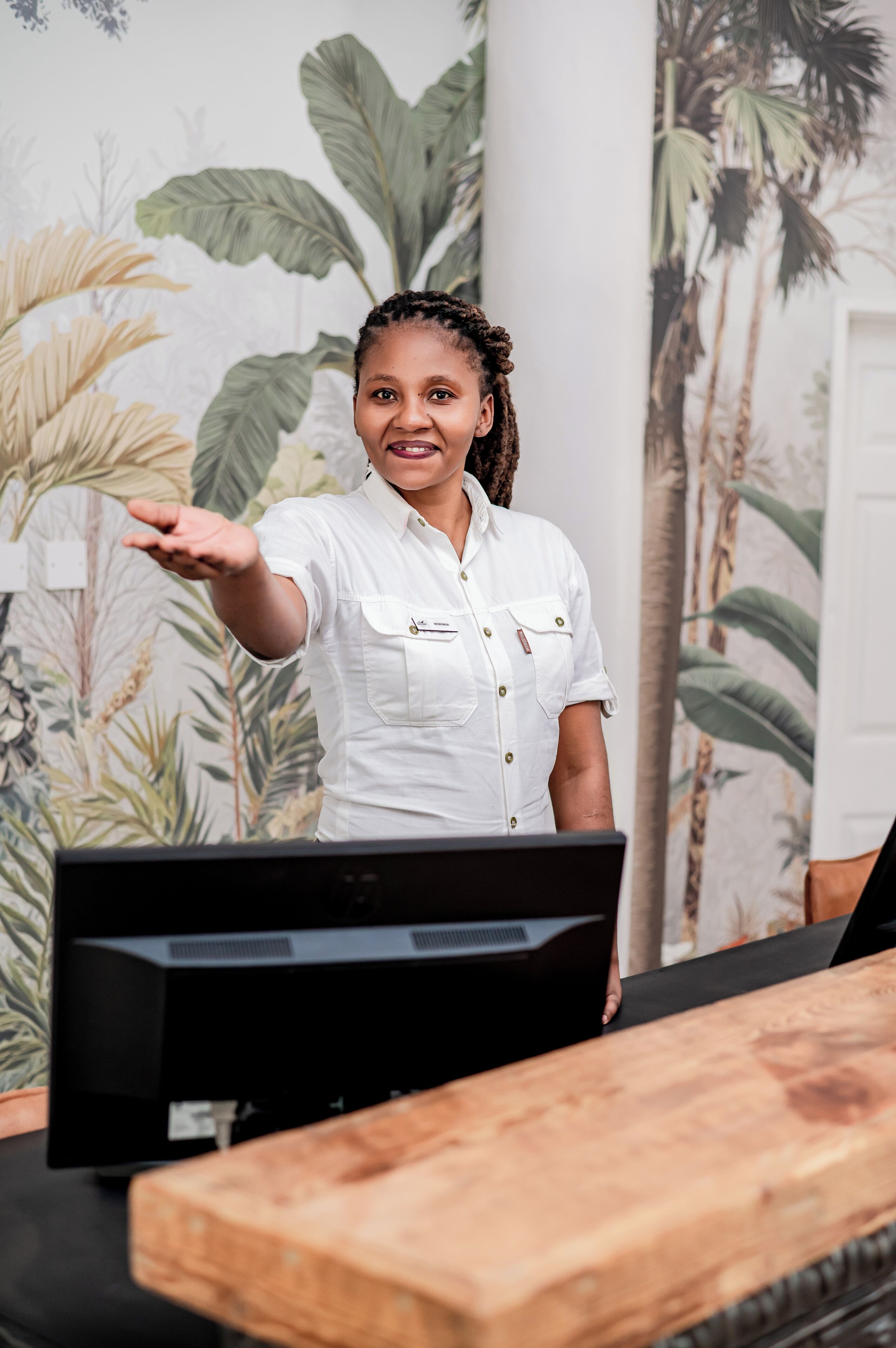 A person standing at a reception desk with a computer monitor in front of them, a wooden counter, and a wall with tropical plant wallpaper.