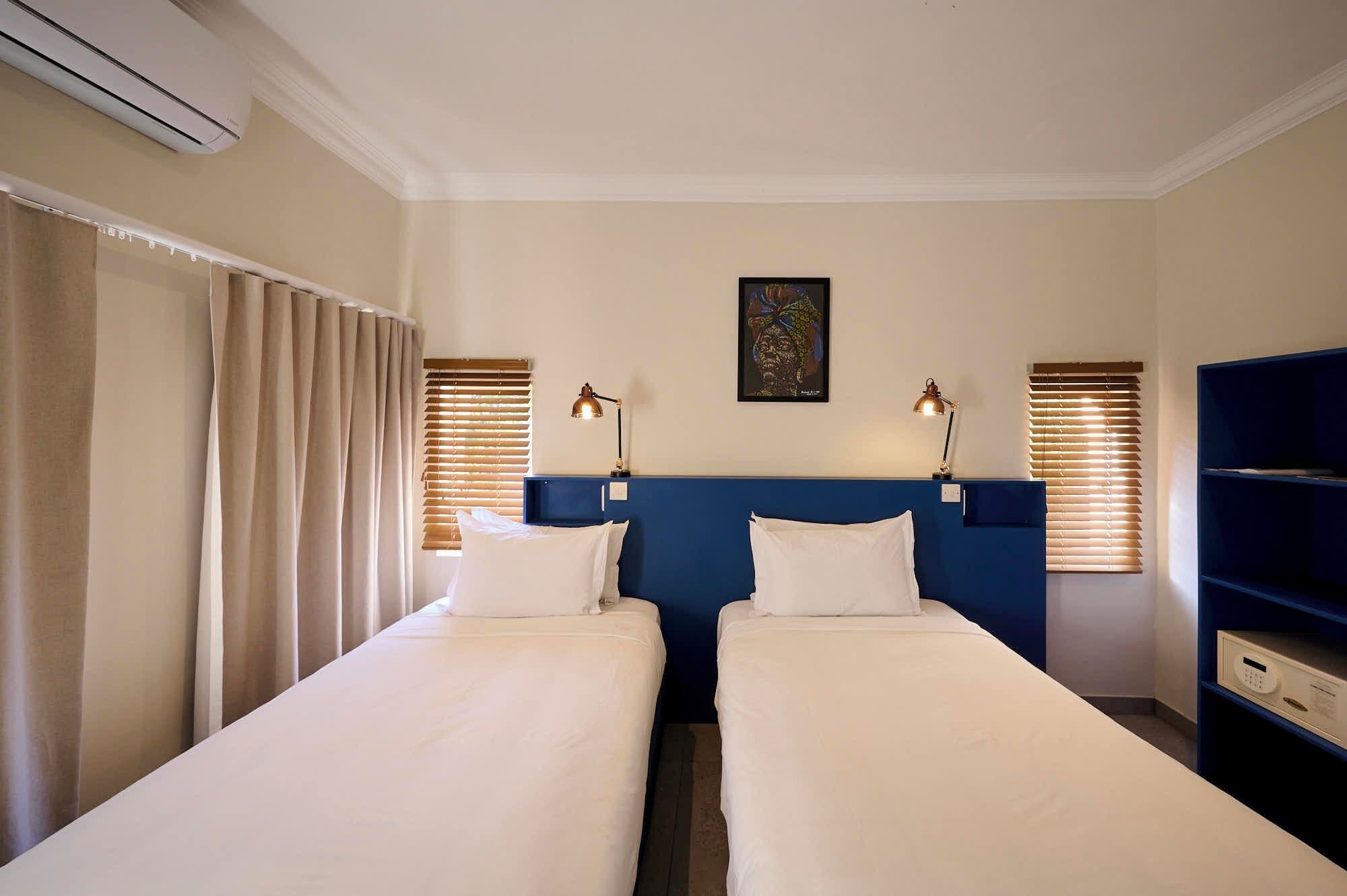 Habitación de hotel con dos camas individuales, cabecera azul, escritorio con silla y una estantería.