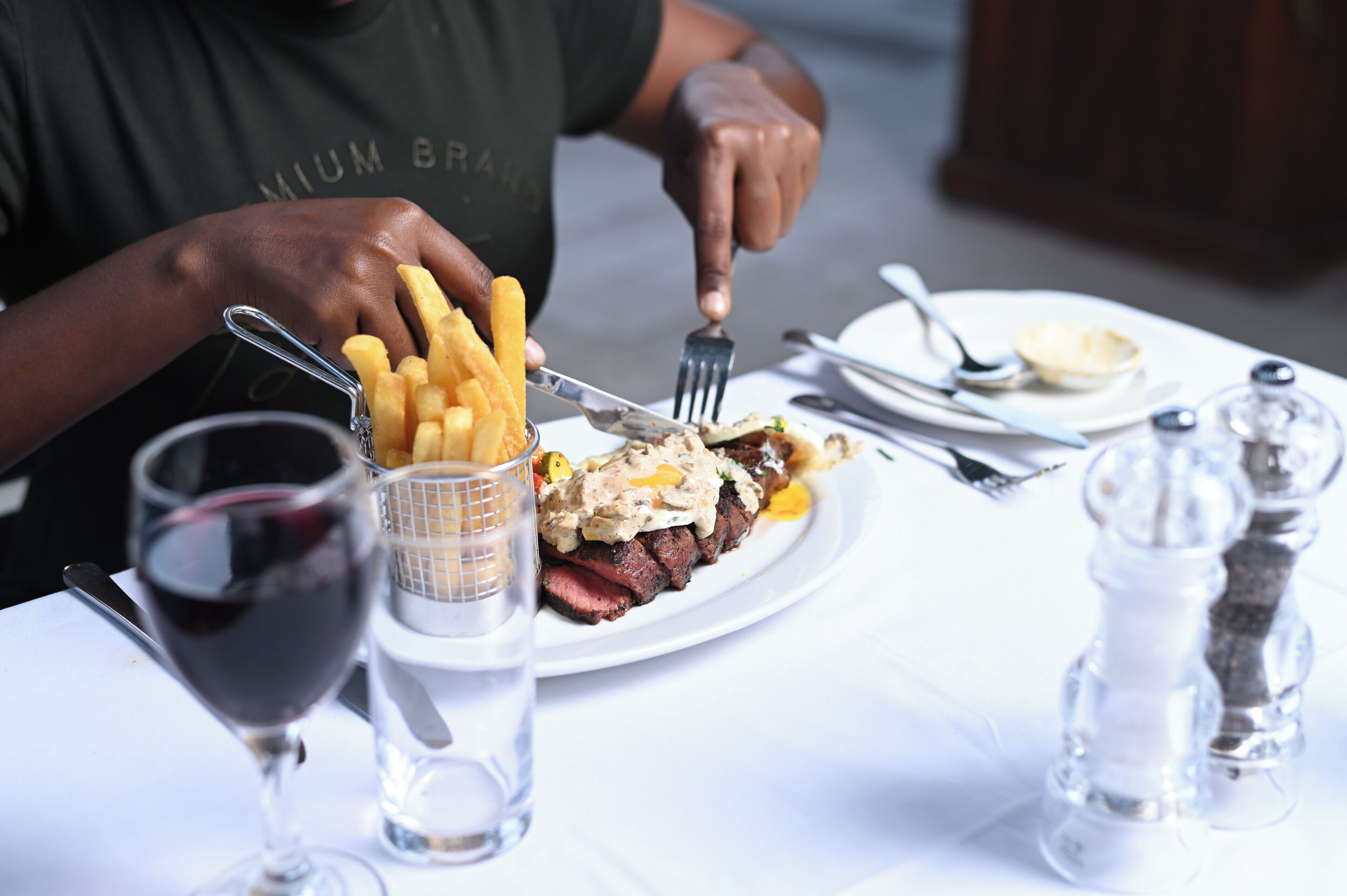 Eine Person isst an einem Tisch, auf dem ein Teller Essen, ein Glas Rotwein und ein Salz- und Pfefferstreuer stehen.
