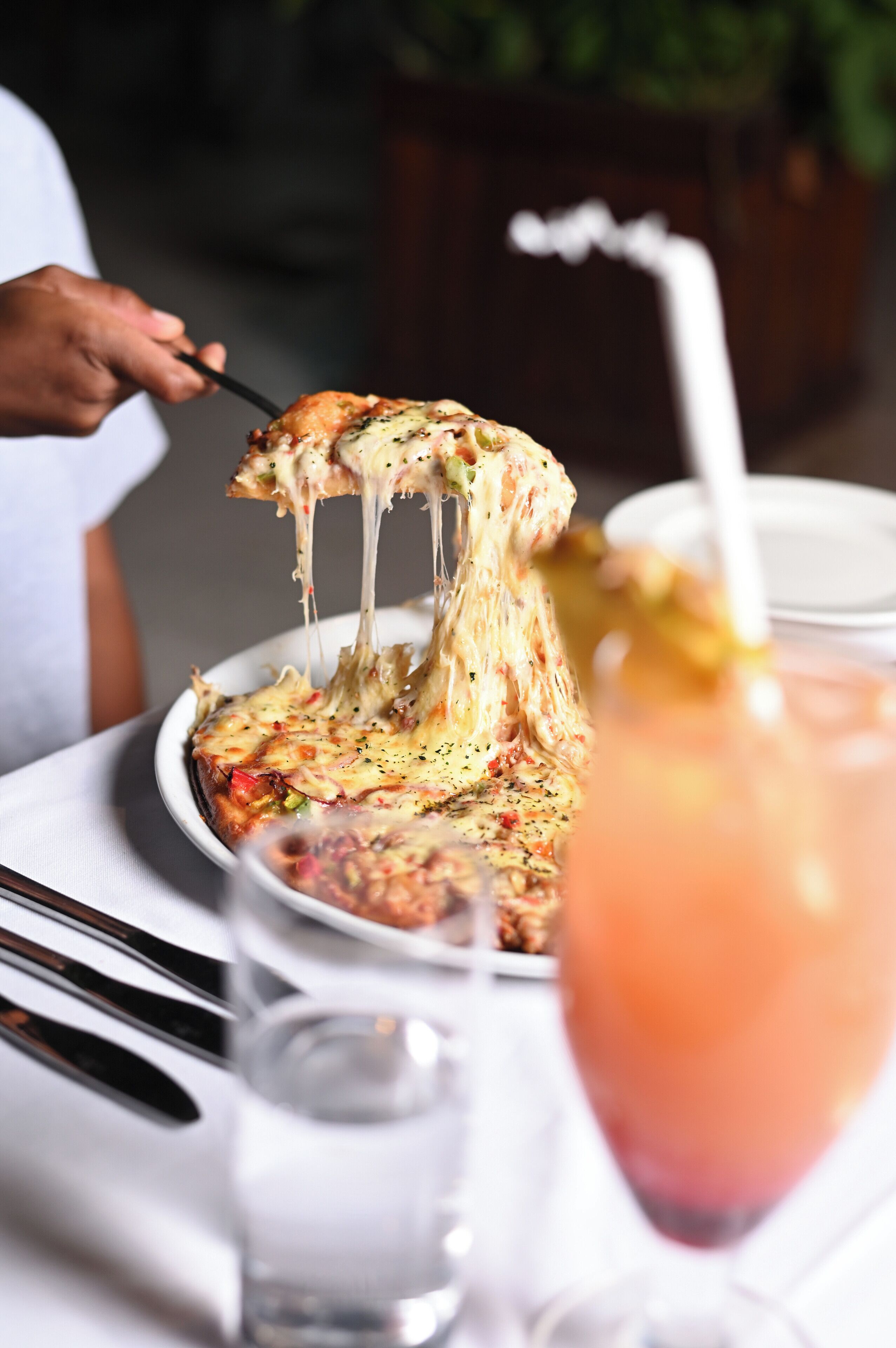 Une pizza à la croûte dorée, recouverte de fromage fondu, est soulevée par une fourchette. À côté, on aperçoit un verre d’eau et un verre à cocktail muni d’une paille.