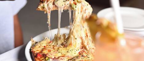 Une pizza à la croûte dorée, recouverte de fromage fondu, est soulevée par une fourchette. À côté, on aperçoit un verre d’eau et un verre à cocktail muni d’une paille.