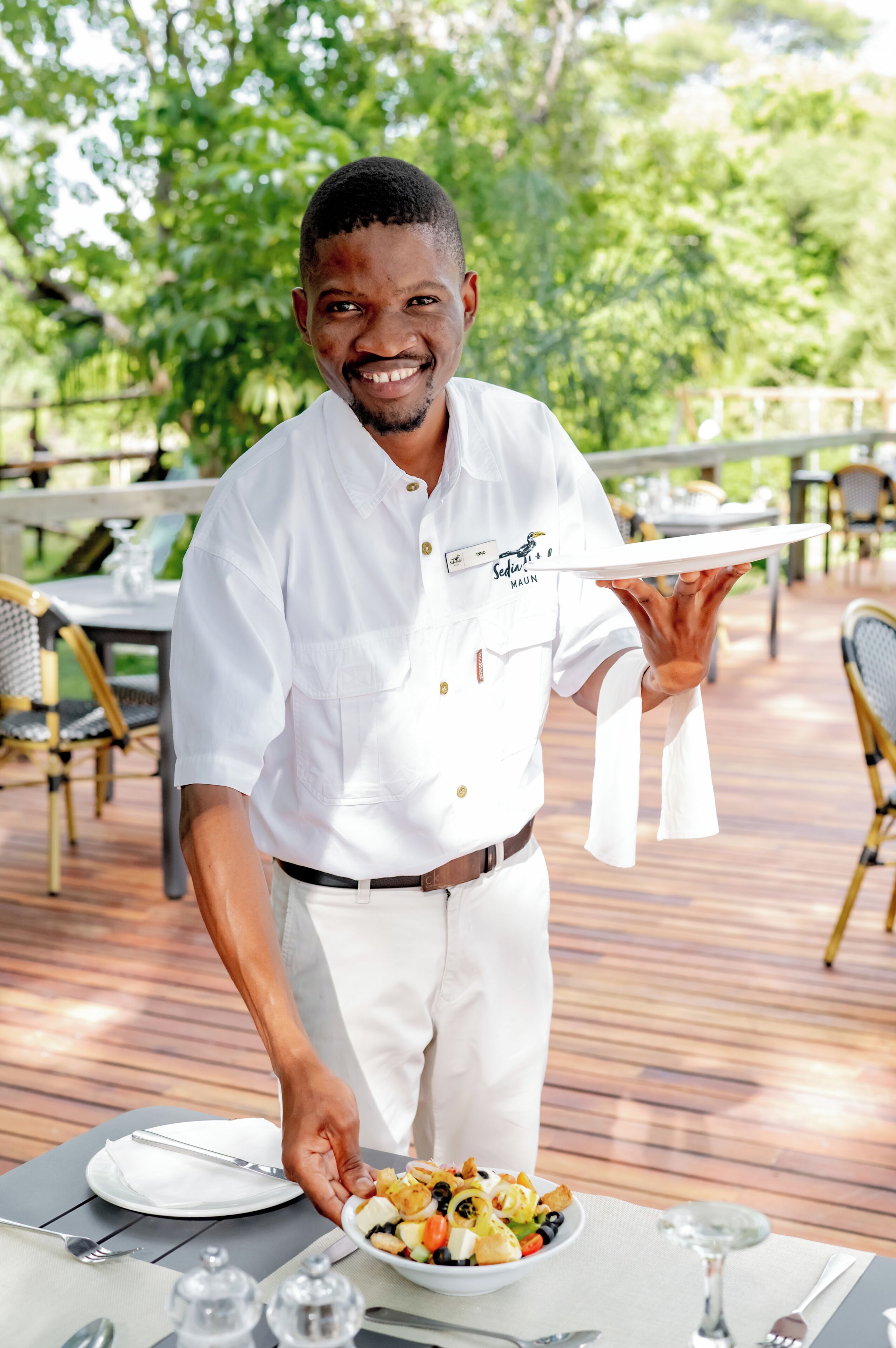A waiter in a white uniform serving a bowl of fruit salad at an outdoor dining table with chairs and a wooden deck.