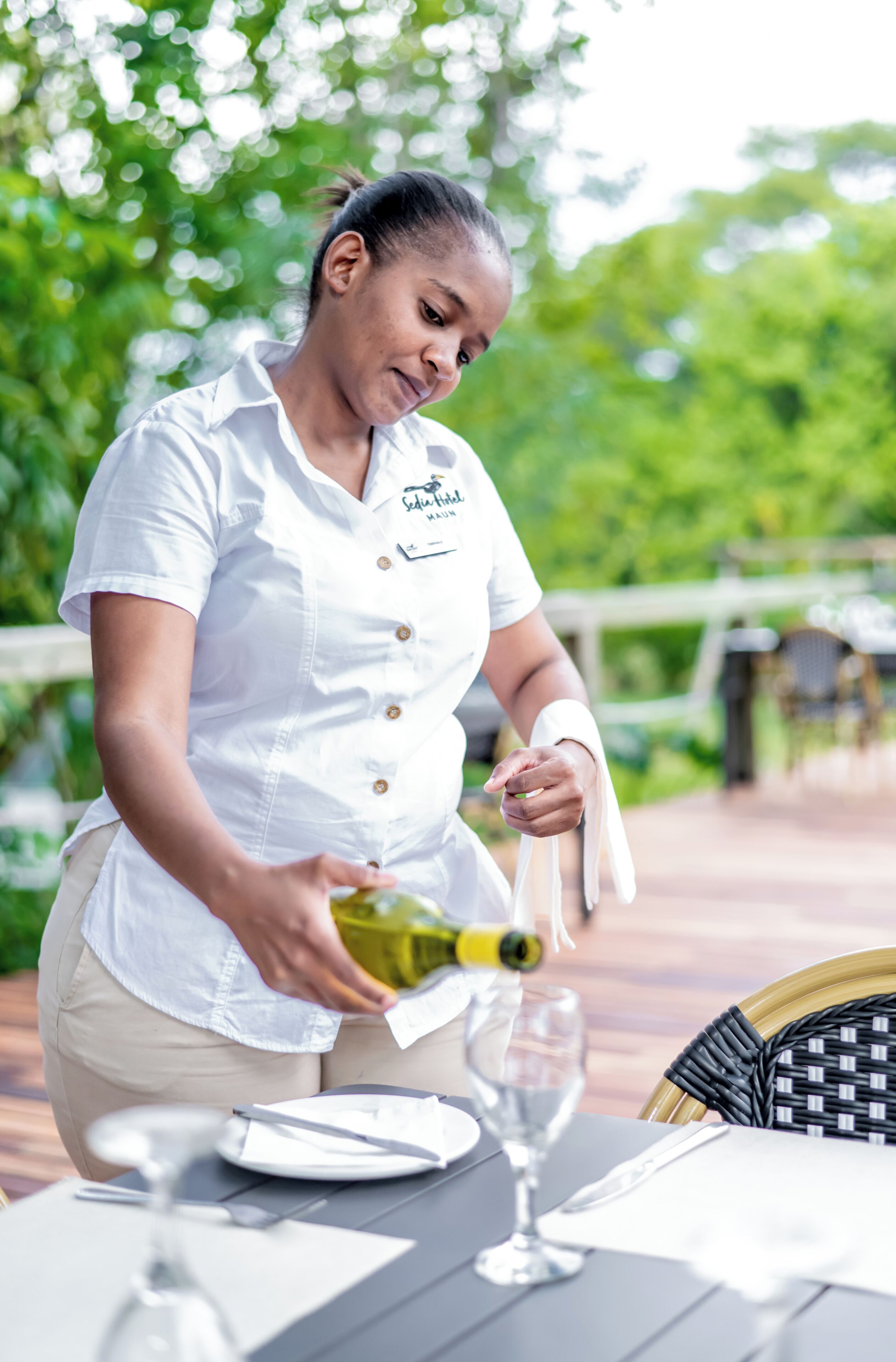 A person pouring a drink into a glass at an outdoor table with a wicker chair and a white tablecloth.