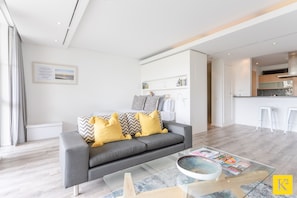 Una sala moderna con un sofá gris, almohadas amarillas y una mesa de centro de vidrio, con una zona de cocina al fondo.