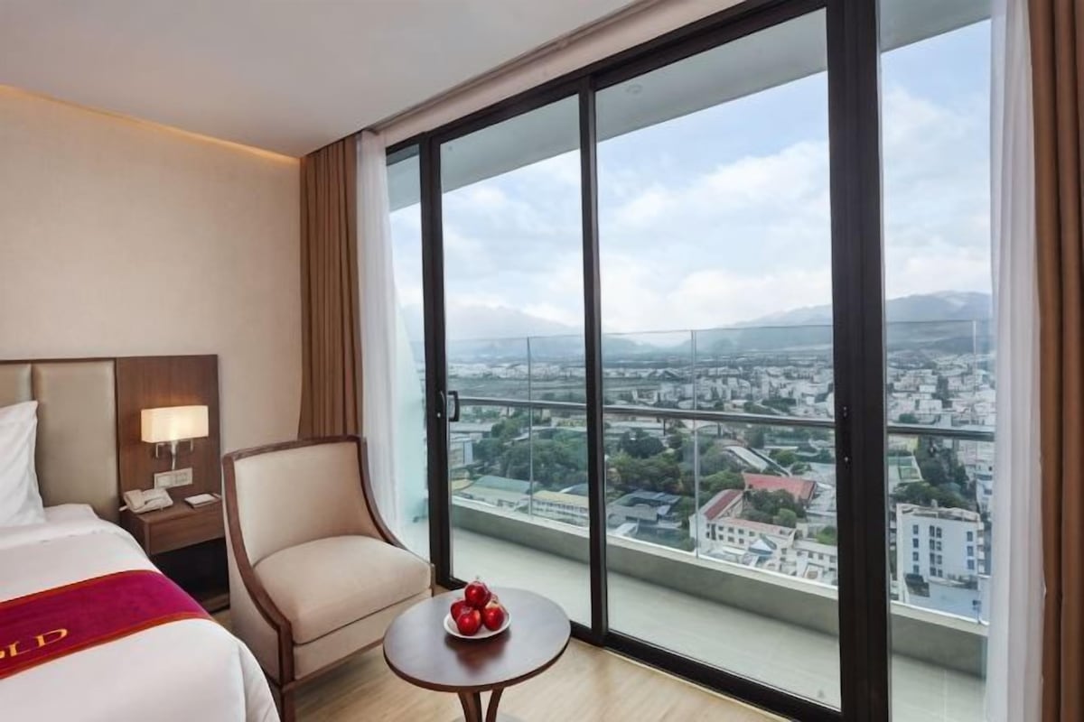 Quarto de hotel com uma cama grande, uma cadeira, uma pequena mesa com frutas e vista para o horizonte urbano através de grandes janelas.