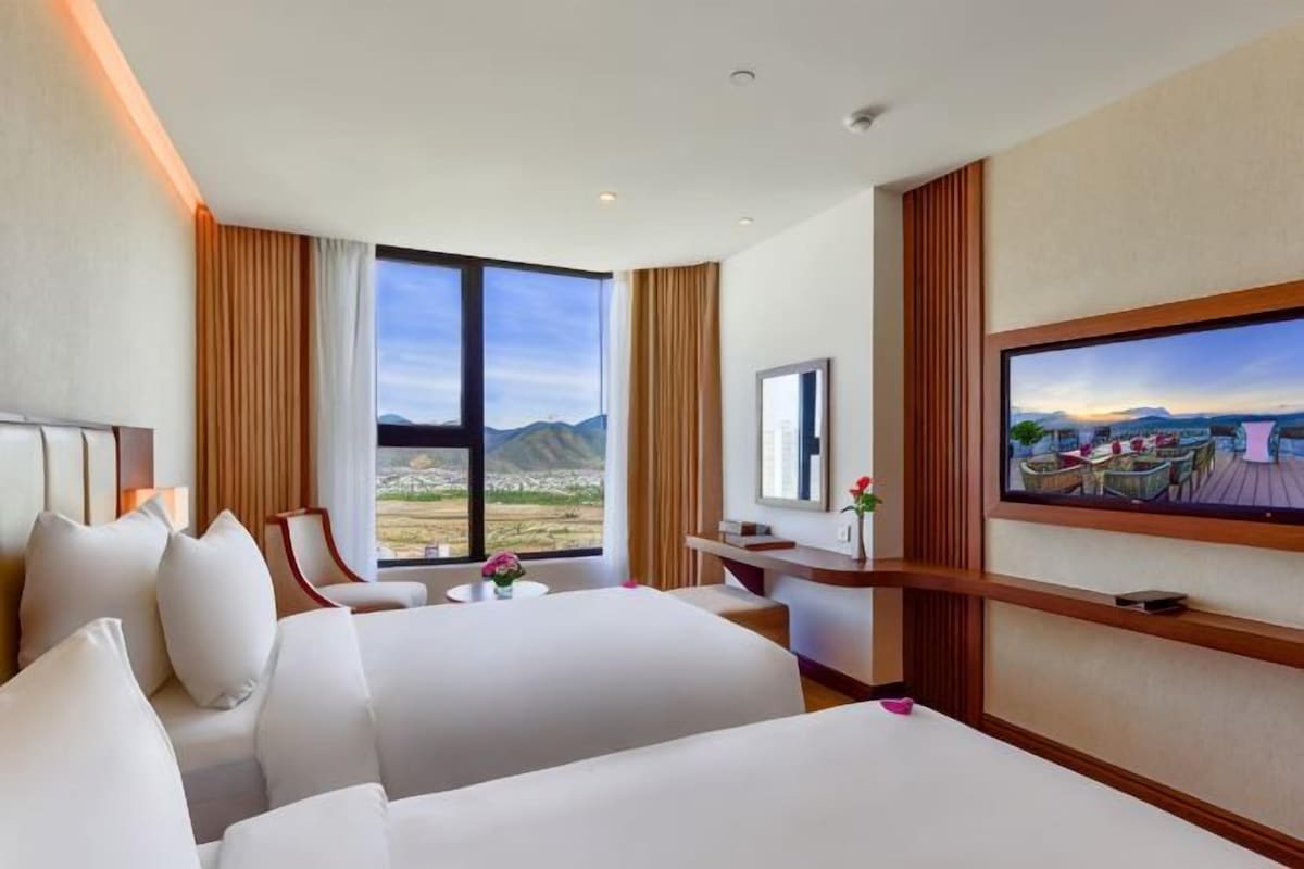 Quarto de hotel com duas camas, uma escrivaninha, televisão e uma grande janela com vista para montanhas.