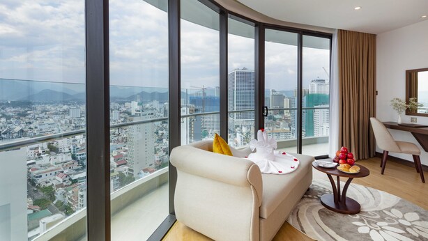 Uma sala de estar moderna com um sofá, uma mesa pequena com uma travessa de fruta, e vista da cidade através de grandes janelas.