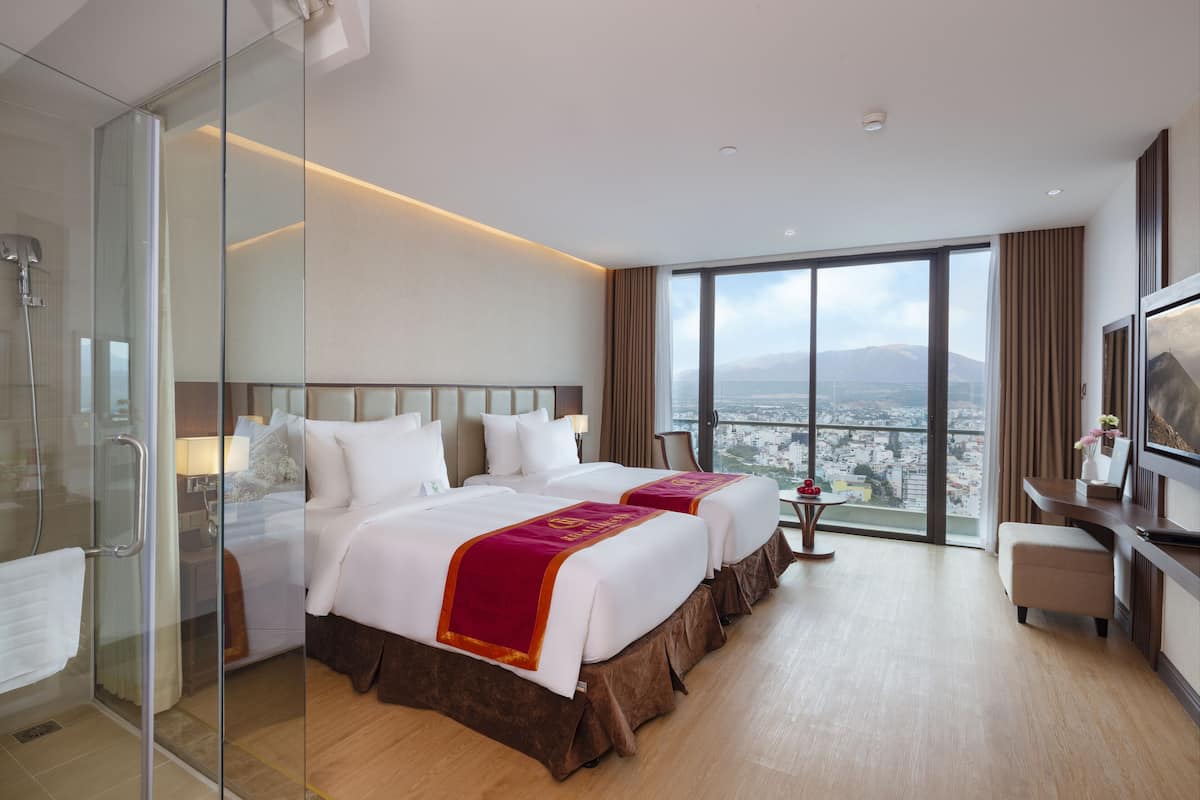 Habitación de hotel con dos camas, cabina de ducha de vidrio, un ventanal con vistas a la ciudad y un escritorio con silla.