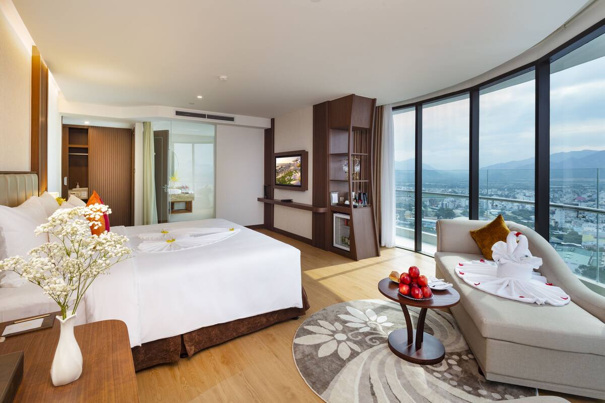 Habitación de hotel con una cama grande, un sofá, una pequeña mesa con fruta, un jarrón con flores, un televisor y un ventanal con vistas a la ciudad.