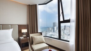 A hotel room with a bed, a chair, a small table with apples, a nightstand with a lamp, and a city view through large windows. - Regalia Gold Hotel (Nha Trang)