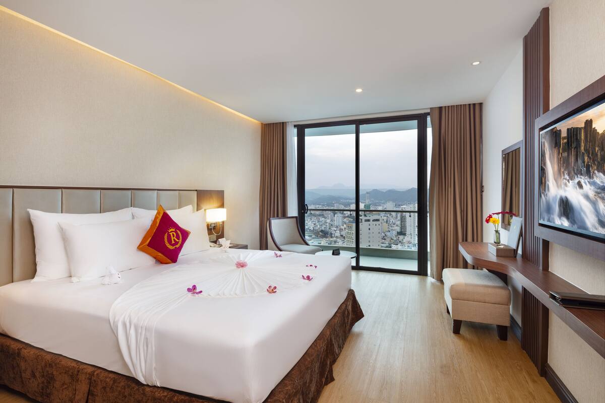 Habitación de hotel con una cama grande, televisión, escritorio y vistas a un paisaje urbano a través de amplias ventanas.