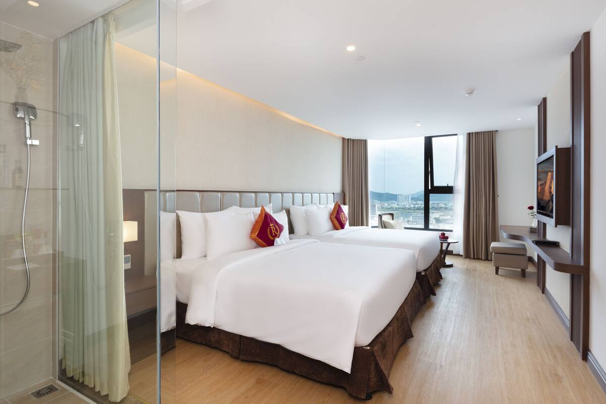 Habitación de hotel con dos camas, ducha, televisión y vista a la ciudad.