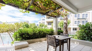 An outdoor patio with a table and chairs, surrounded by greenery and a view of a building complex. - 001 Altmore on the Waterfront Marina (Cape Town)
