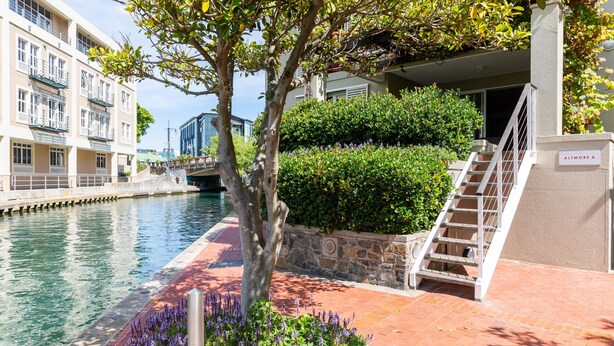 A building with a staircase leading to a balcony, a tree, and a water canal. - 001 Altmore on the Waterfront Marina (Cape Town)