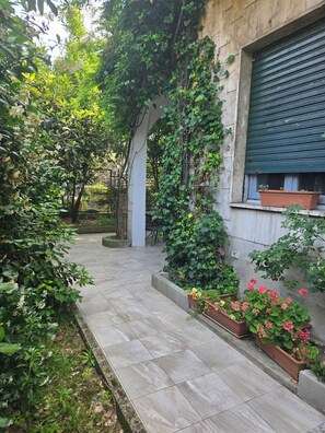 Une cour dallée, entourée de verdure et bordée d’un bâtiment doté d’une fenêtre et d’un volet fermé.