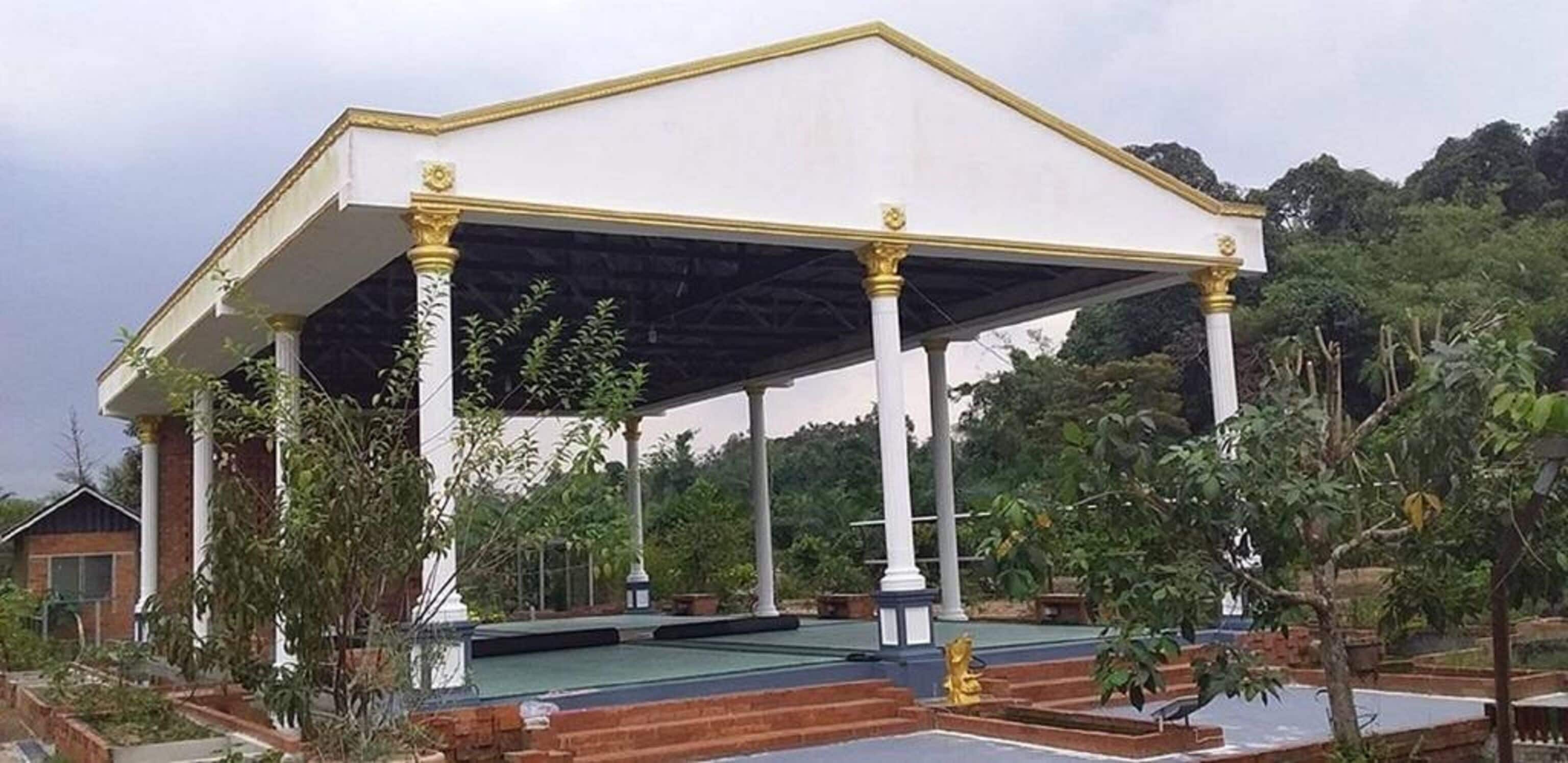 Um pavilhão coberto com colunas brancas e teto plano, rodeado por vegetação e uma piscina.