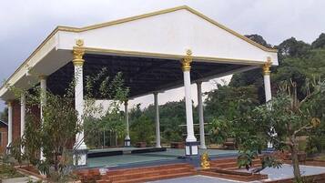 Um pavilhão coberto com colunas brancas e teto plano, rodeado por vegetação e uma piscina.