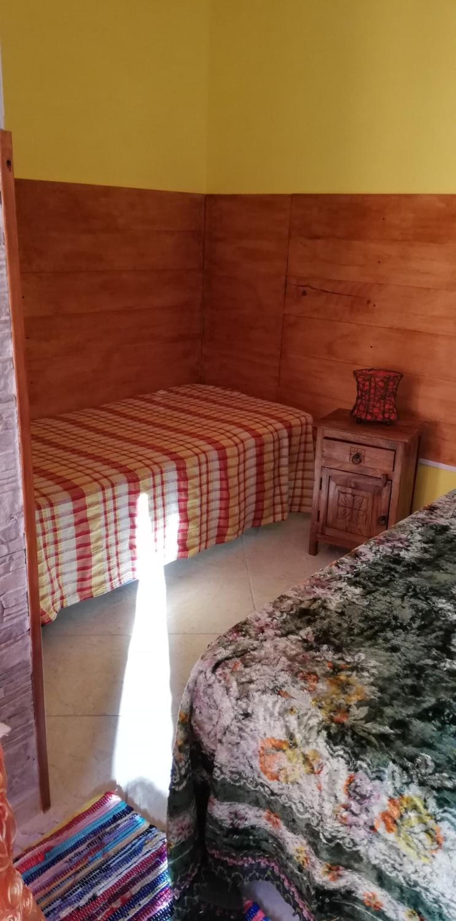 A bedroom with a wooden headboard, a bed with a checkered bedspread, a nightstand with a vase, and a colorful striped rug.
