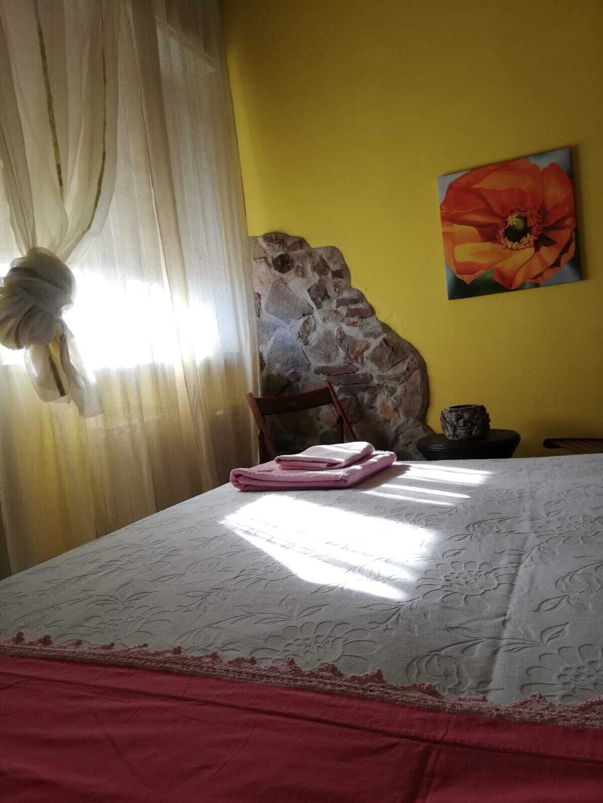 A bedroom with a bed, a stone wall, a painting of a flower, and a window with curtains.
