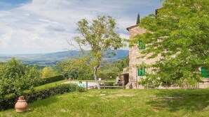 Um edifício rústico de pedra com piscina, rodeado por vegetação exuberante e uma vista panorâmica de colinas distantes.