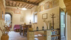 Um interior rústico com vigas de madeira aparente, lareira de pedra, área de jantar com mesa e cadeiras e uma estante cheia de livros.