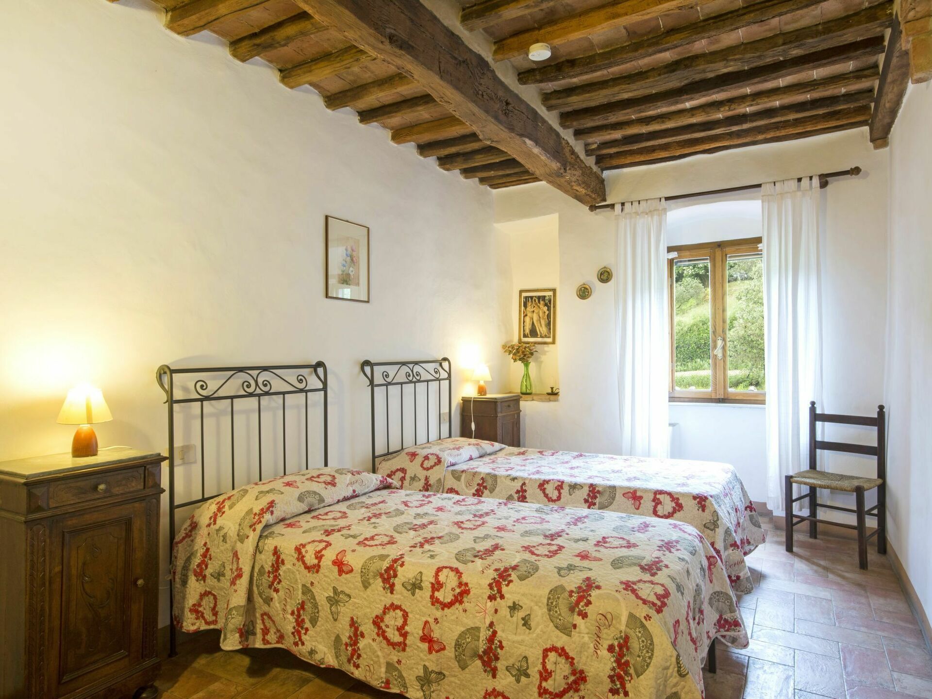 Camera da letto con due letti, testiere in legno, un comodino con una lampada, una sedia e una finestra con tende.