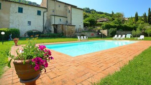 Uma piscina com cadeiras de descanso brancas, um cachepot de terracota com flores e um edifício rústico ao fundo.