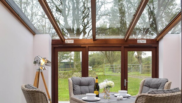 Un coin repas aménagé avec des meubles en osier, une table dressée pour deux personnes et une vue sur le jardin grâce à de grandes portes vitrées.