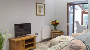 Un salon avec un téléviseur posé sur un meuble en bois, un vase rempli de fleurs sur une petite table et un canapé recouvert d’une couverture.