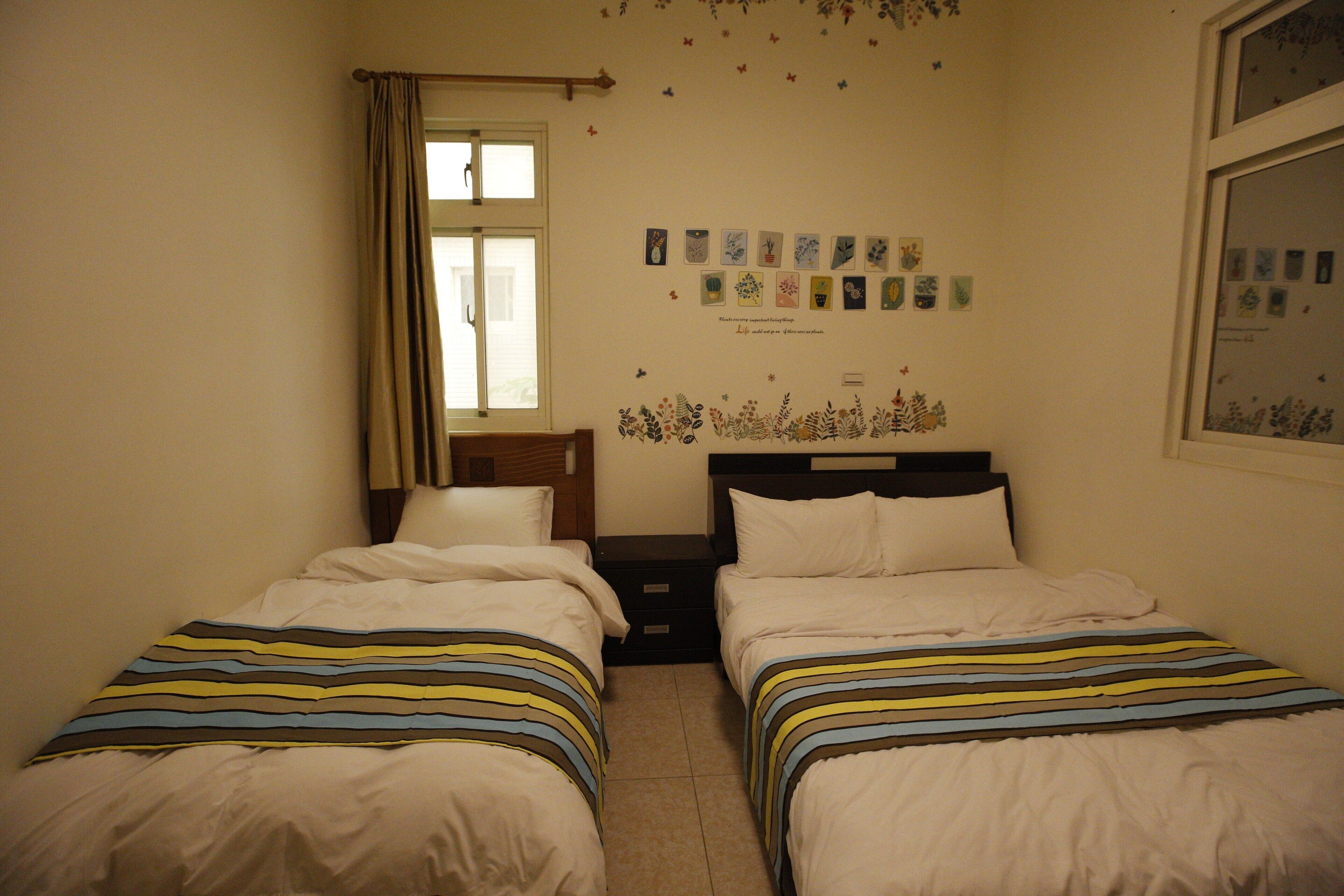 Un dormitorio con dos camas, una ventana con cortinas, una mesita de noche y una pared decorada con fotos pequeñas.