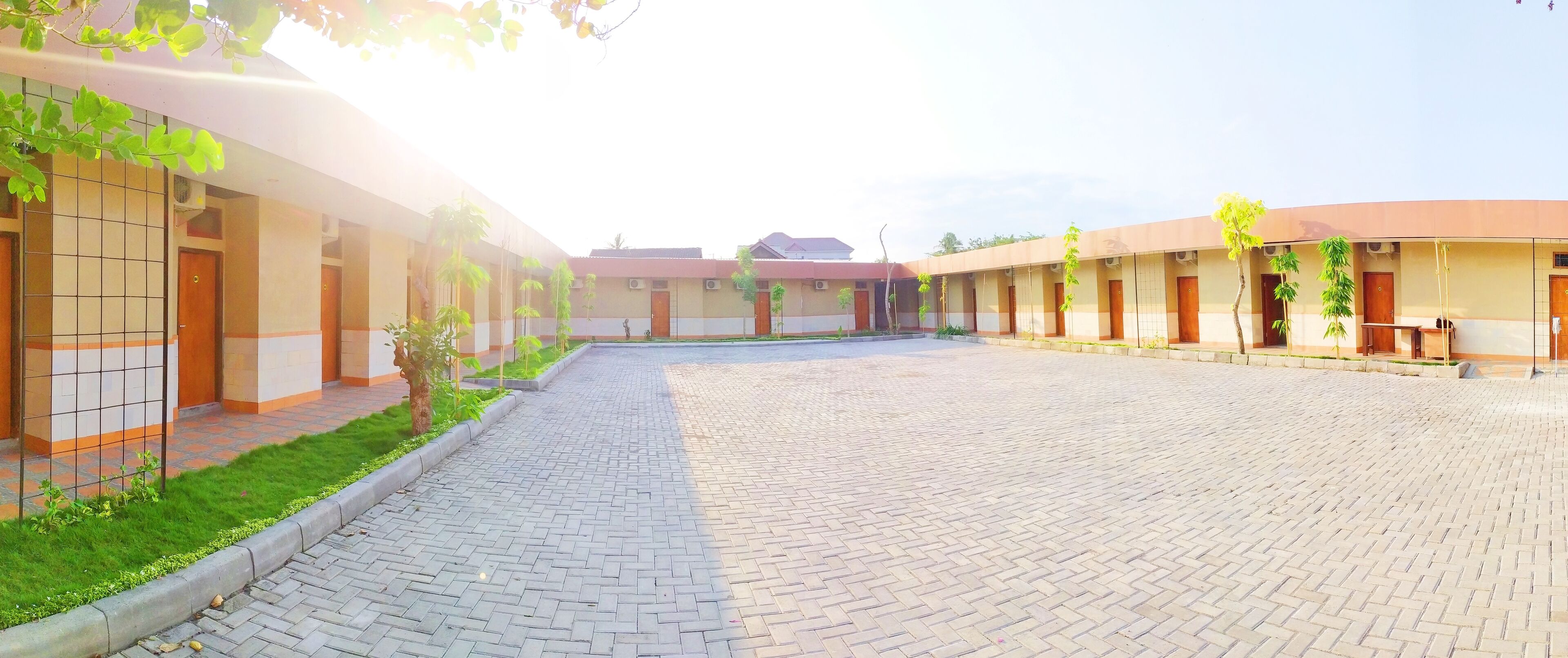 A courtyard with a paved walkway, surrounded by buildings with doors and windows, and small trees along the edges.