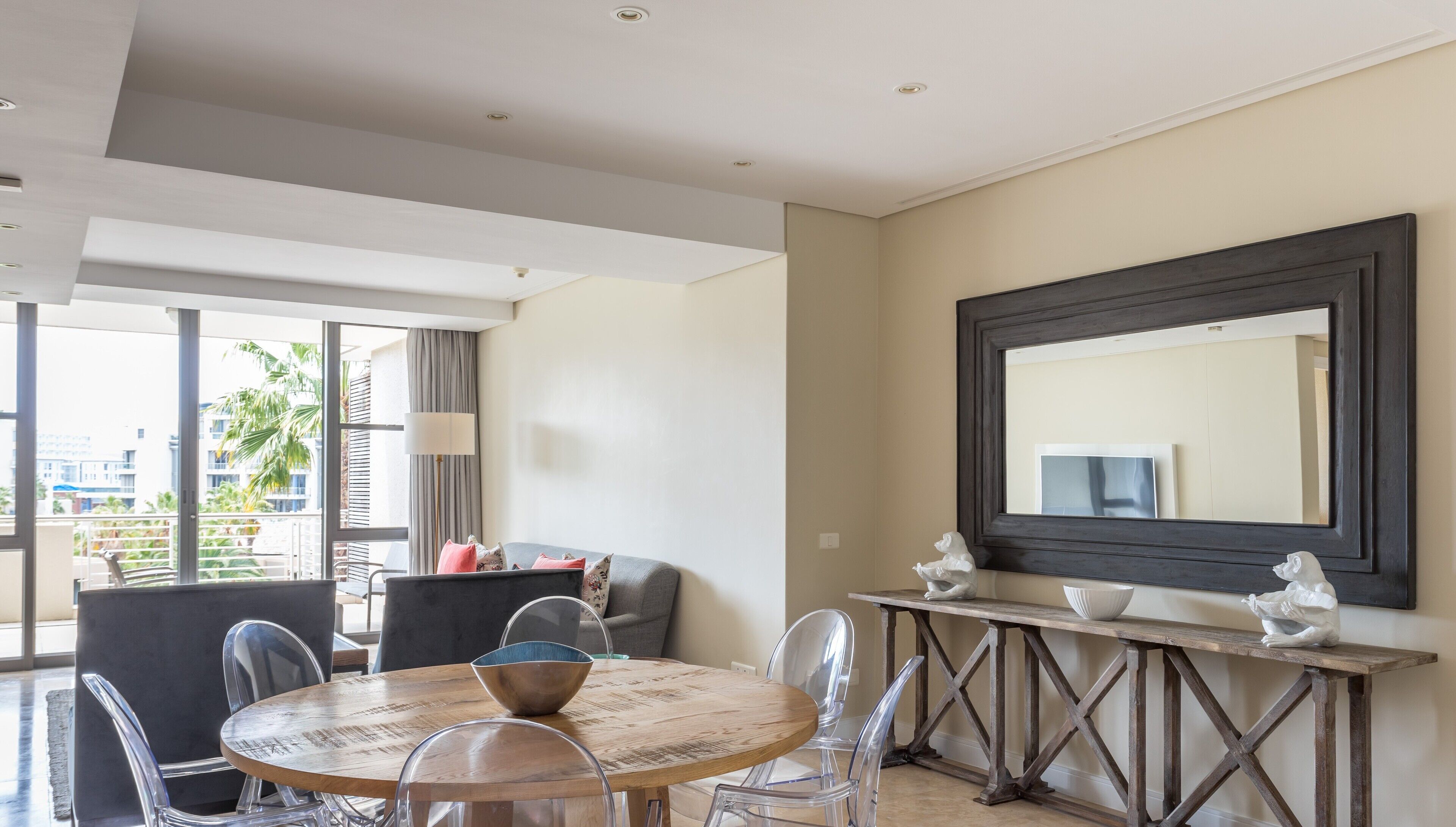Quarto de hotel moderno com mesa de jantar redonda, cadeiras transparentes, sofá cinza, um espelho grande e uma varanda com vista para palmeiras.