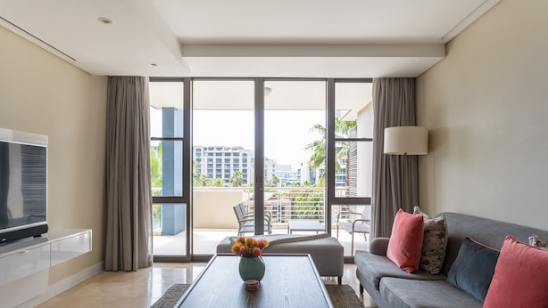 A modern hotel room with a flat-screen TV, a sofa with decorative pillows, a coffee table with a vase of flowers, and a large sliding glass door leading to a balcony with a city view. - 308 Juliette on the Waterfront Marina (Cape Town)