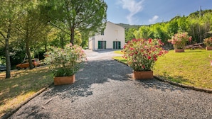 A gravel driveway leading to a white building with green shutters, surrounded by lush greenery and flowering plants. - Nice apartment in villa for 17 people with pool, TV and pets allowed (Rosignano Solvay-castiglioncello)