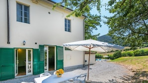 A white building with green shutters, a gravel patio, a table with chairs, and a large umbrella. - Nice apartment in villa for 17 people with pool, TV and pets allowed (Rosignano Solvay-castiglioncello)