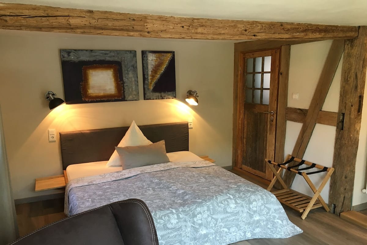 Een slaapkamer met een houten plafond, een bed met een bloemenbedekking, twee wandlampen en een houten ladder.