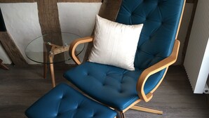 Een blauwe gestoffeerde fauteuil met een passende poef, geplaatst naast een glazen tafel met houten frame.