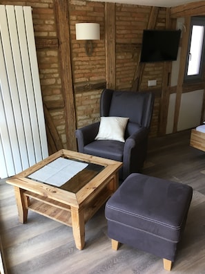A room with a wooden table, a cushioned ottoman, a leather chair, a wall-mounted lamp, and a television on a brick wall. - Schmuckkästle am Hafen (Wiesbaden)
