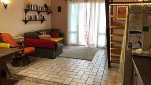 Ein gemütliches Wohnzimmer mit Sofa, Sessel und Esstisch. Ein Bücherregal beherbergt Bücher und Dekoration. Ein Kühlschrank mit Magneten und Zetteln ist zu sehen.