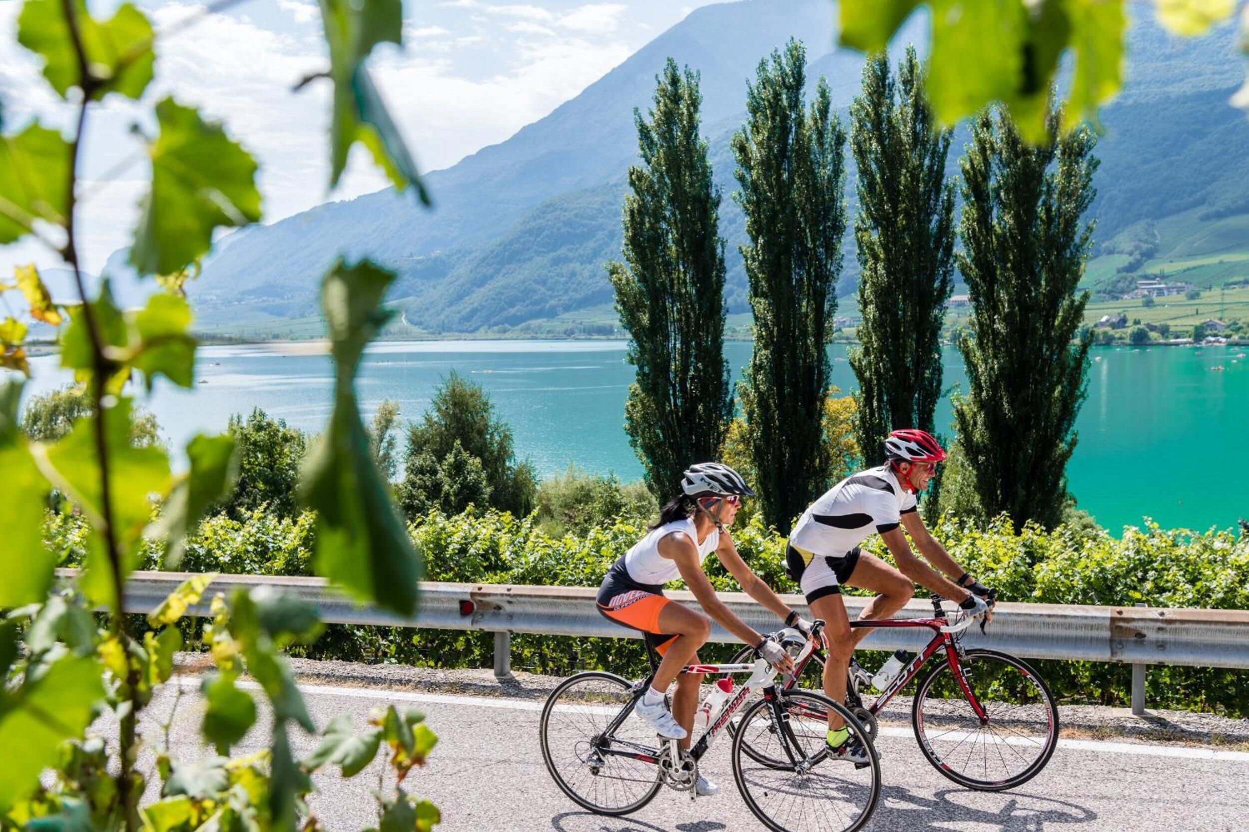 Deux cyclistes sur une route, avec un lac et des montagnes en arrière-plan.