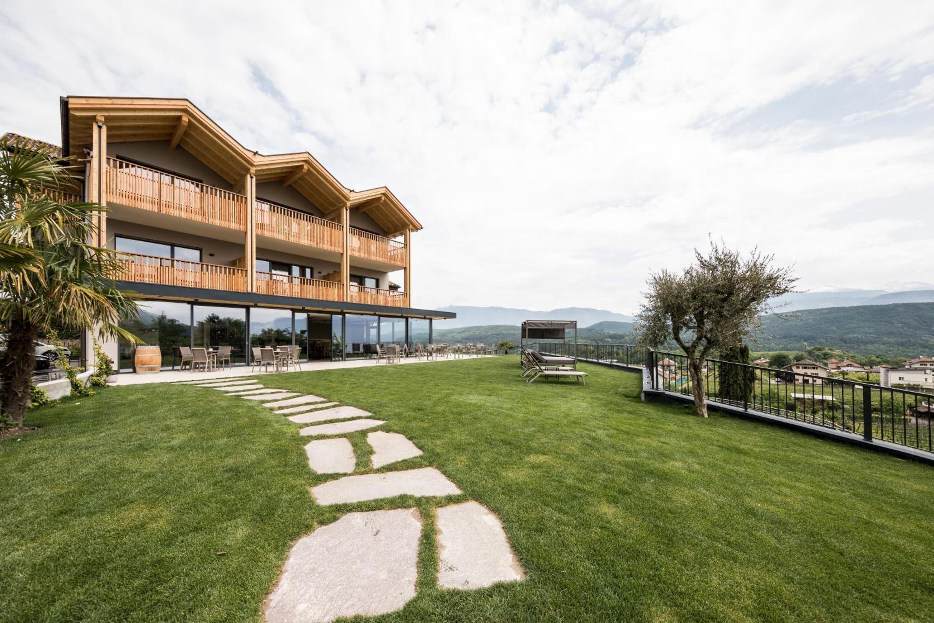 Un edificio moderno de dos pisos con un balcón de madera, un camino de piedra y vistas del paisaje circundante.