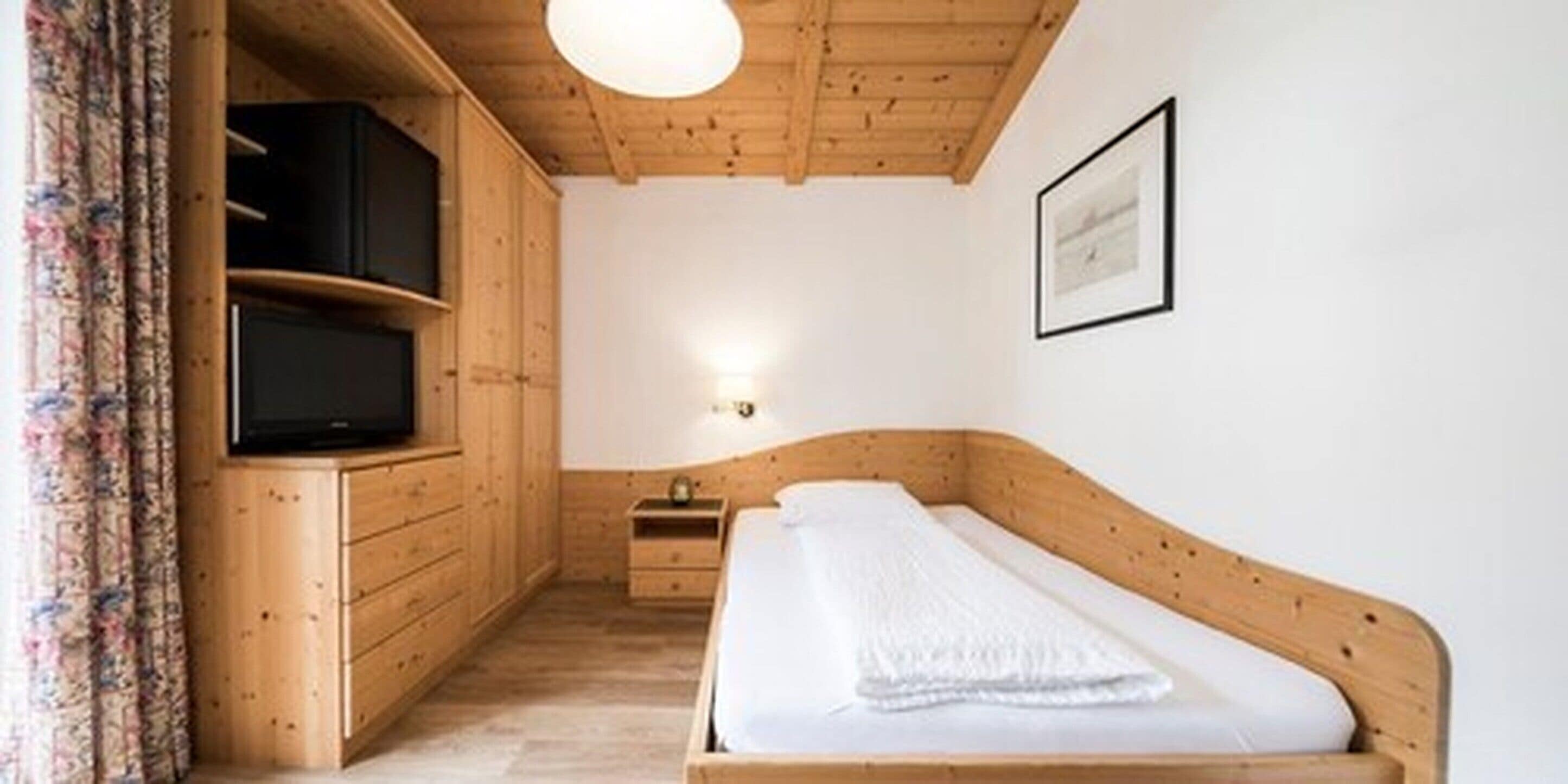 Habitació de fusta amb un llit, una televisió sobre un moble de fusta, una tauleta de nit i una làmpada penjada a la paret.