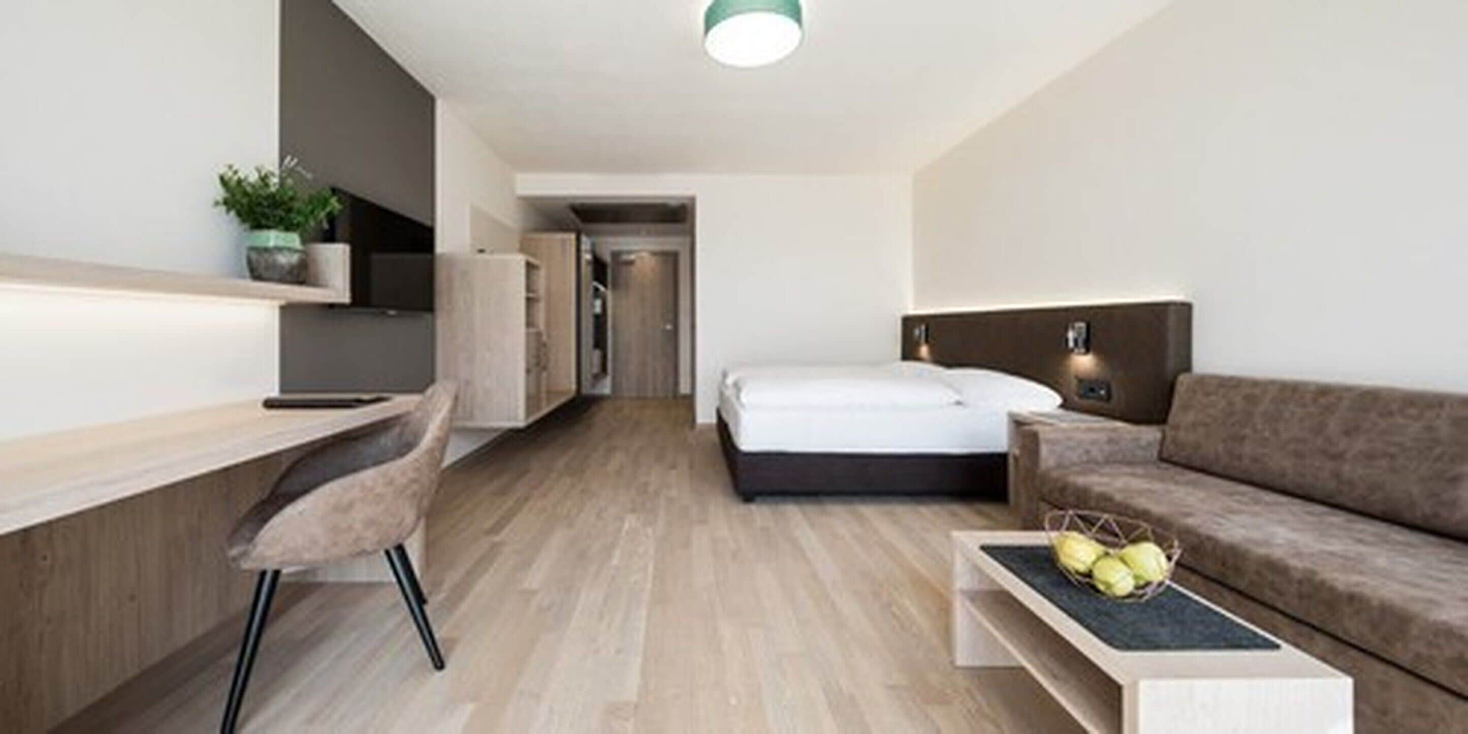 Quarto de hotel moderno com chão de madeira, uma cama, sofá, escrivaninha e uma cadeira.