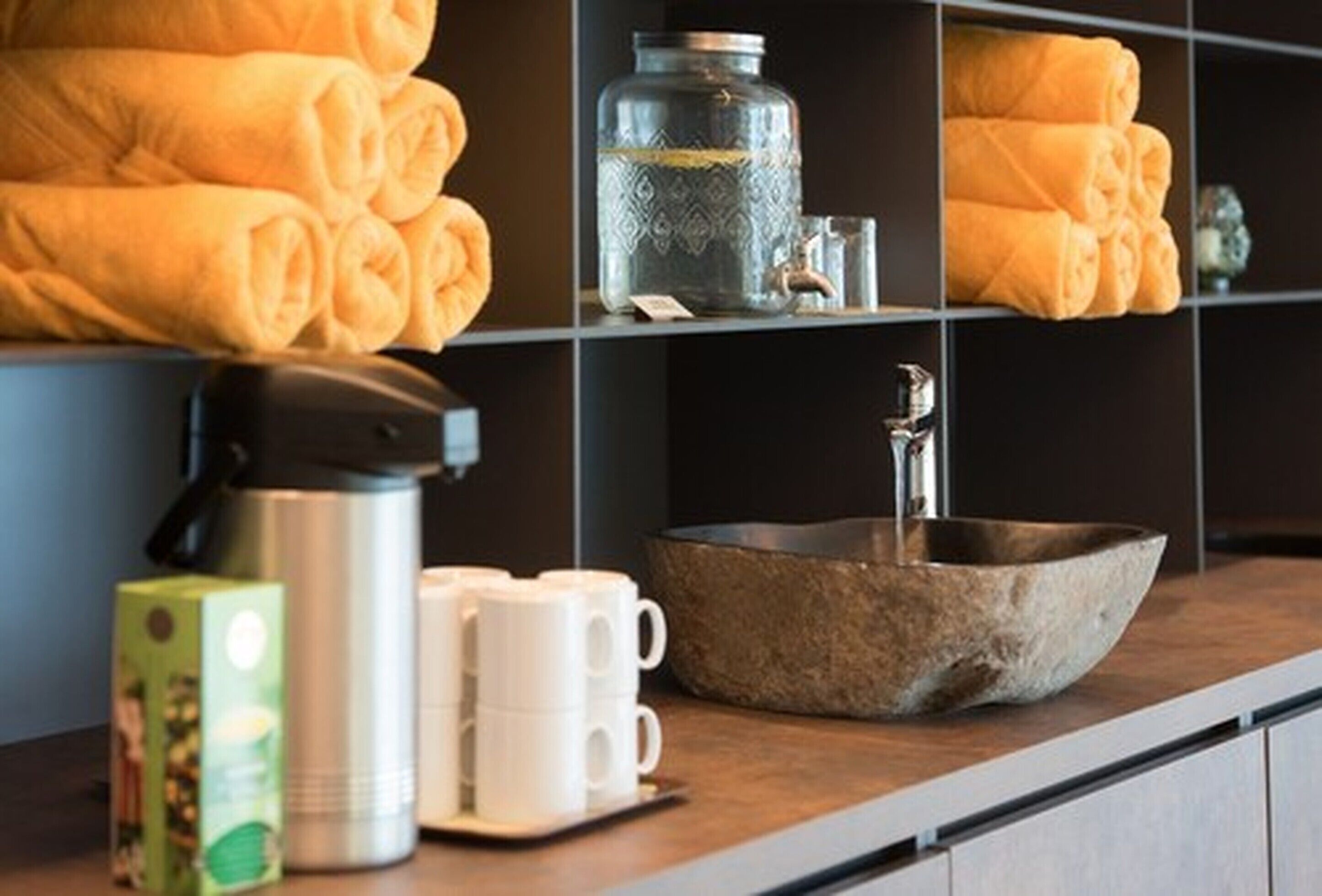 Une chambre d’hôtel avec un comptoir en bois, une cafetière en métal, des tasses blanches, un bol en pierre et des serviettes soigneusement pliées, rangées sur des étagères.