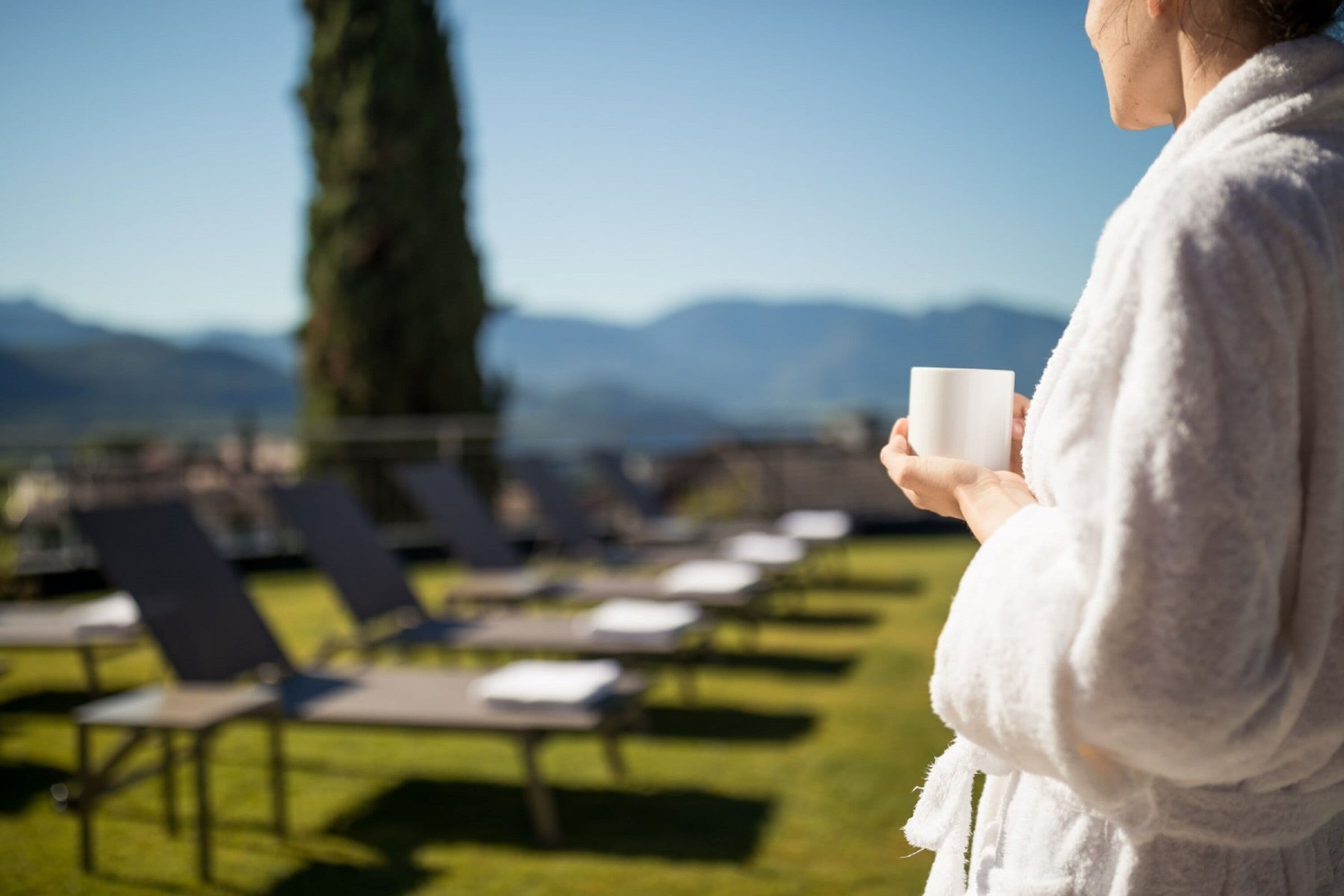 A person in a white robe holding a white cup, standing on a grassy area with lounge chairs and a view of mountains in the background.