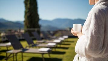 A person in a white robe holding a white cup, standing on a grassy area with lounge chairs and a view of mountains in the background.