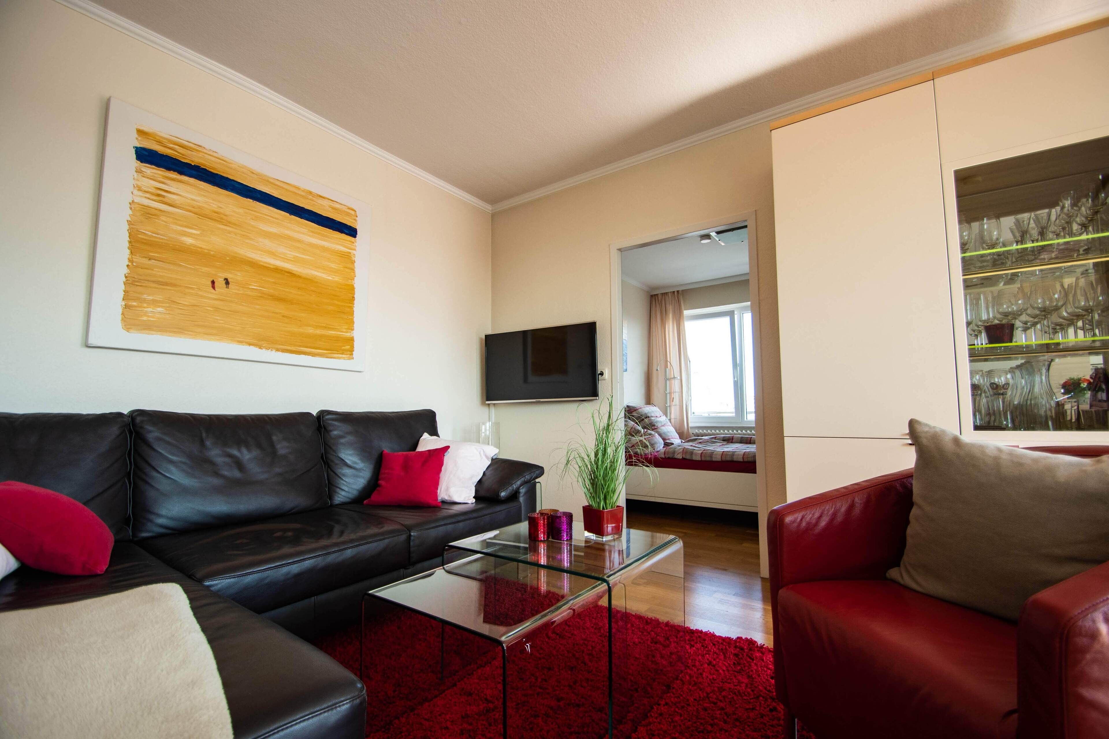 Una sala de estar moderna con un sofá de cuero negro, cojines rojos, una mesa de centro de vidrio y una alfombra roja. Un televisor de pantalla plana está montado en la pared, y hay una planta sobre la mesa de centro.
