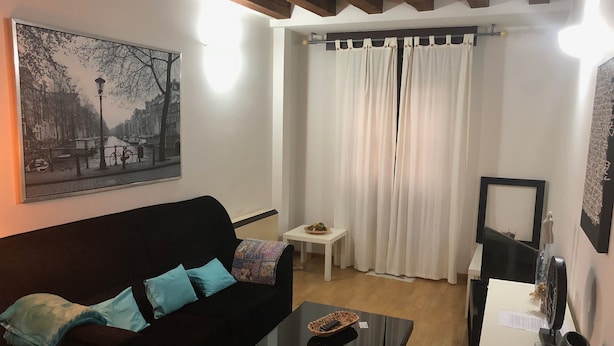A living room with a black sofa, a glass coffee table, a small white side table, and a painting of a cityscape. - Beautiful duplex in unbeatable location (Segovia)