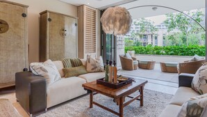 Un salon moderne avec un canapé, une table basse et des fauteuils en osier. Une grande fenêtre offre une vue sur la verdure et les bâtiments.