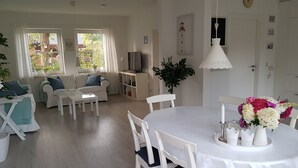 A bright and airy living room with a dining area, featuring a white table, chairs, and a TV on a stand. - **** NEW **** Ferienhaus Meerzeit Eckernförde **** NEW ****  (Eckernförde)